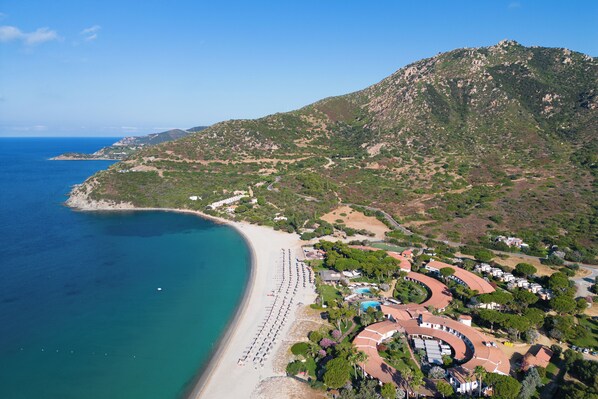 An aerial view of a coastal resort with a long sandy beach, a mountainous backdrop, and a cluster of buildings with curved roofs. - Hotel Residence Cormoran (Villasimius)