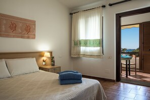A hotel room with a bed, a nightstand, a lamp, a window with curtains, and a view of the outside. - Hotel Residence Cormoran (Villasimius)