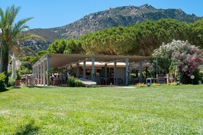An outdoor seating area with tables and chairs, a covered structure, and a mountain backdrop. - Hotel Residence Cormoran (Villasimius)