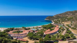 An aerial view of a coastal resort with a curved building, a beach, and a mountainous backdrop.