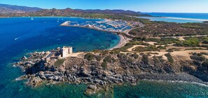 An aerial view of a coastal town with a marina, rocky shoreline, and a tower. - Hotel Residence Cormoran (Villasimius)