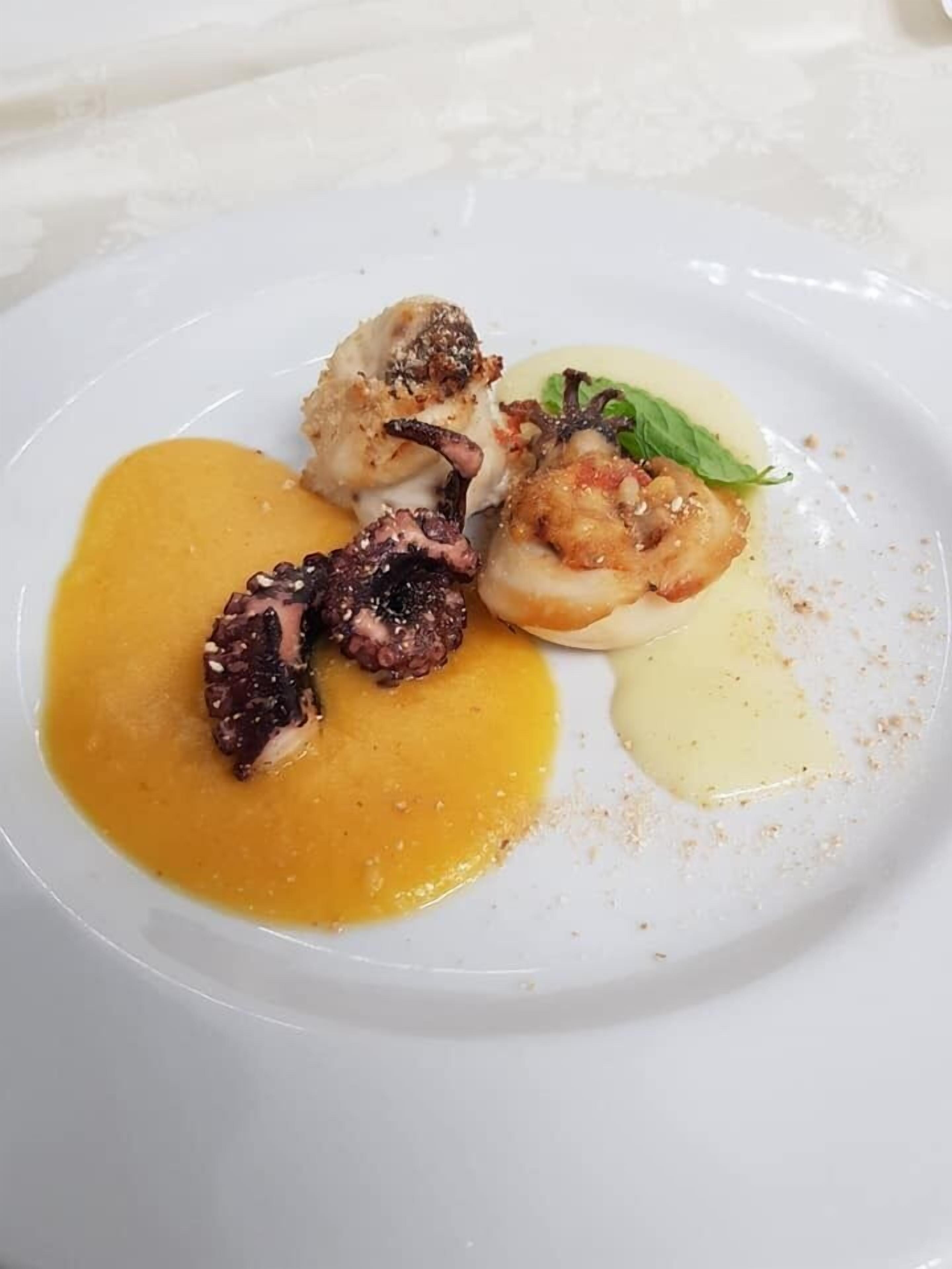 Un plato blanco con un guiso de pulpo, un vieira y un adorno verde sobre un fondo blanco.