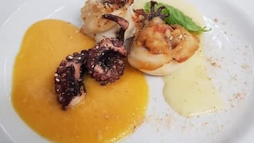 Un plato blanco con un guiso de pulpo, un vieira y un adorno verde sobre un fondo blanco.