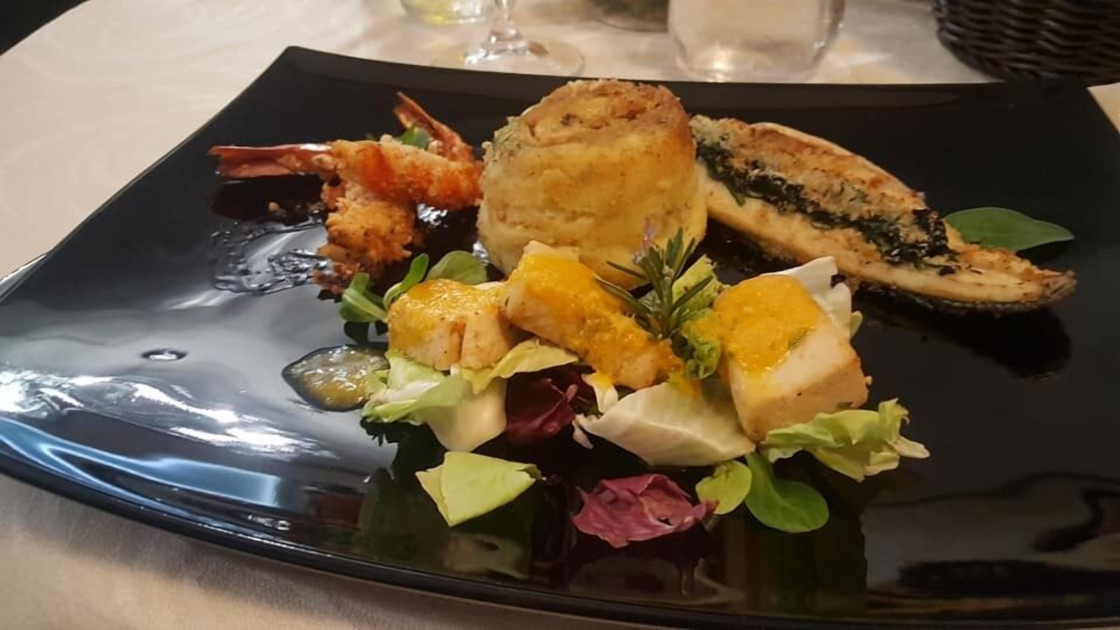 Un plato negro con diversos platos de mariscos, incluyendo camarones, pescado y un preparado empanizado, acompañado de una ensalada y un recipiente pequeño con salsa.