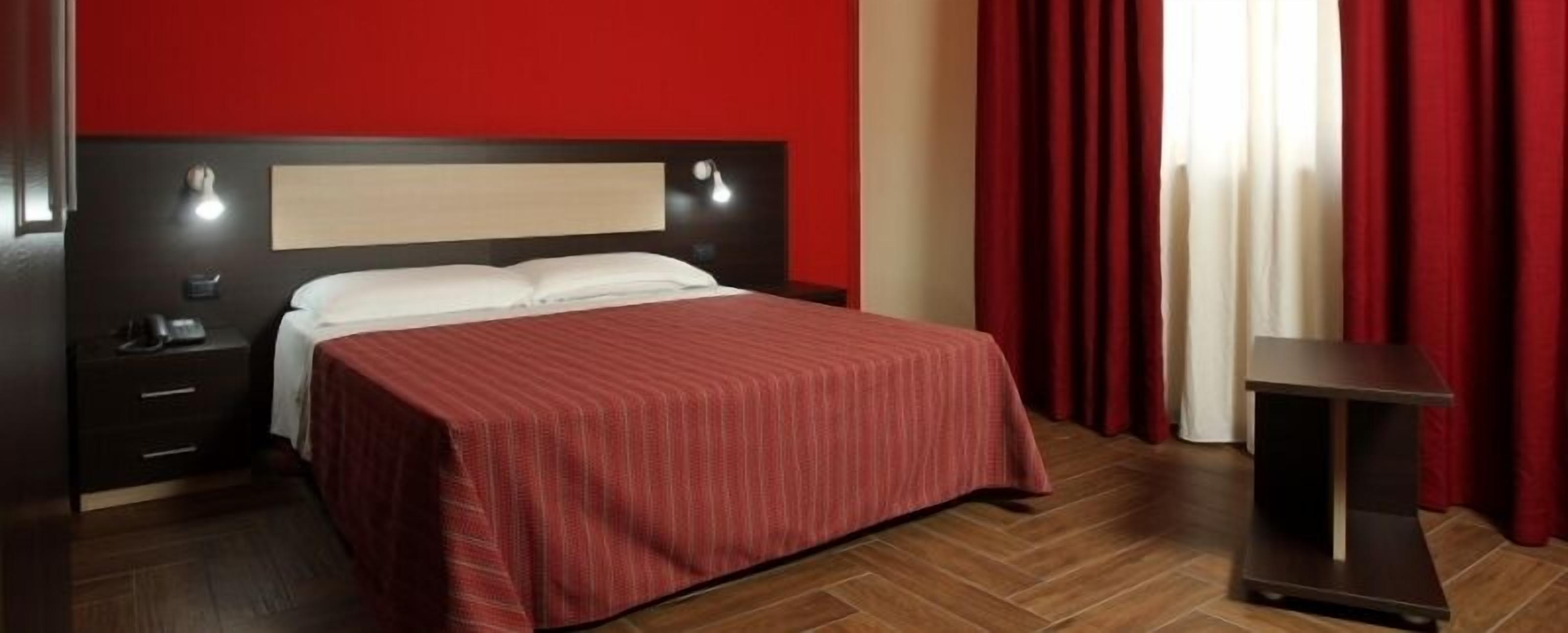 A hotel room with a bed, a nightstand, a desk, and red curtains.