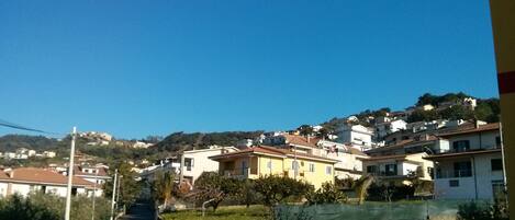 Un barrio residencial con casas en una ladera, cielo despejado y una calle con cerramiento y vegetación.