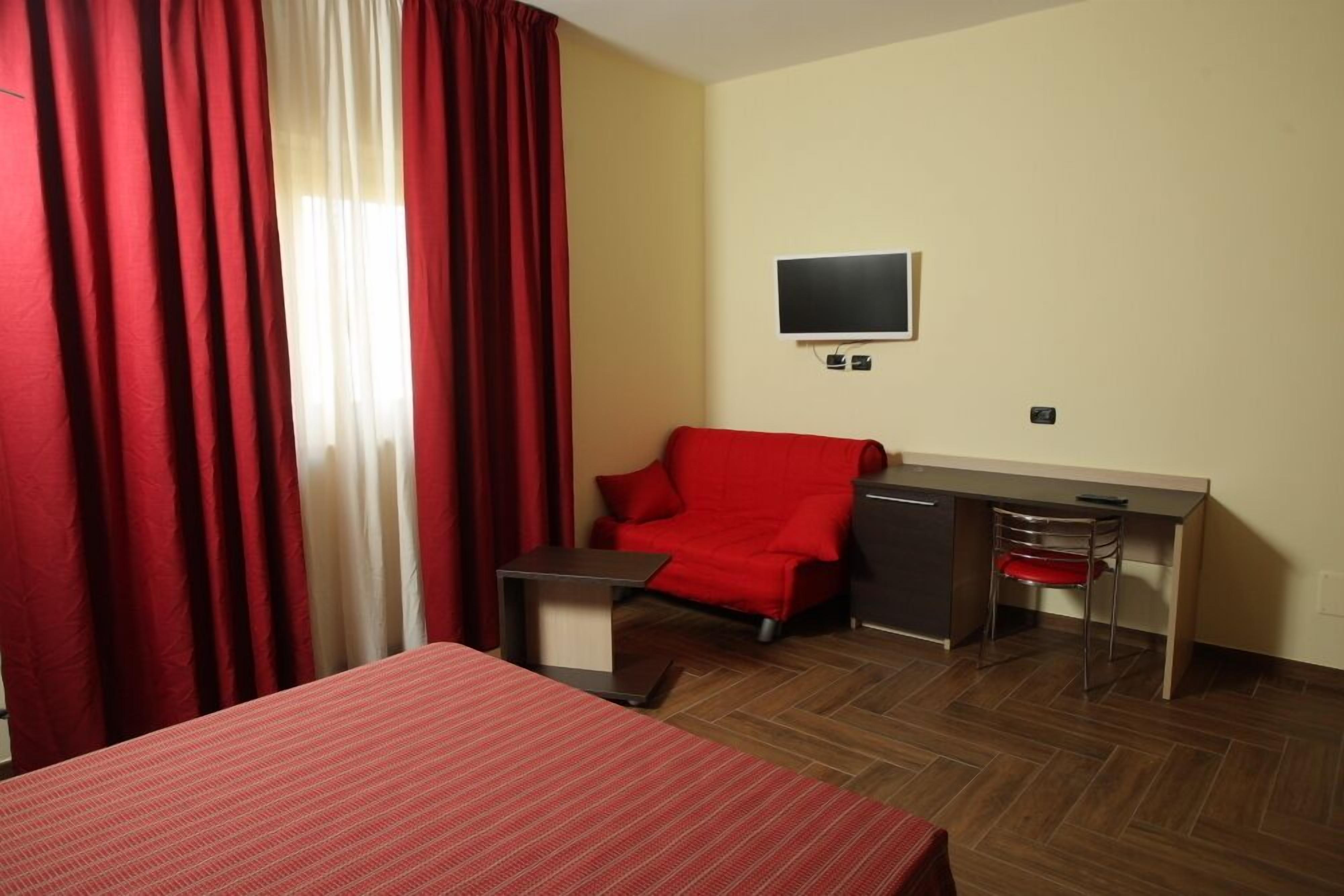 Habitación de hotel con cama, escritorio, silla, televisor y ventana con cortinas rojas.