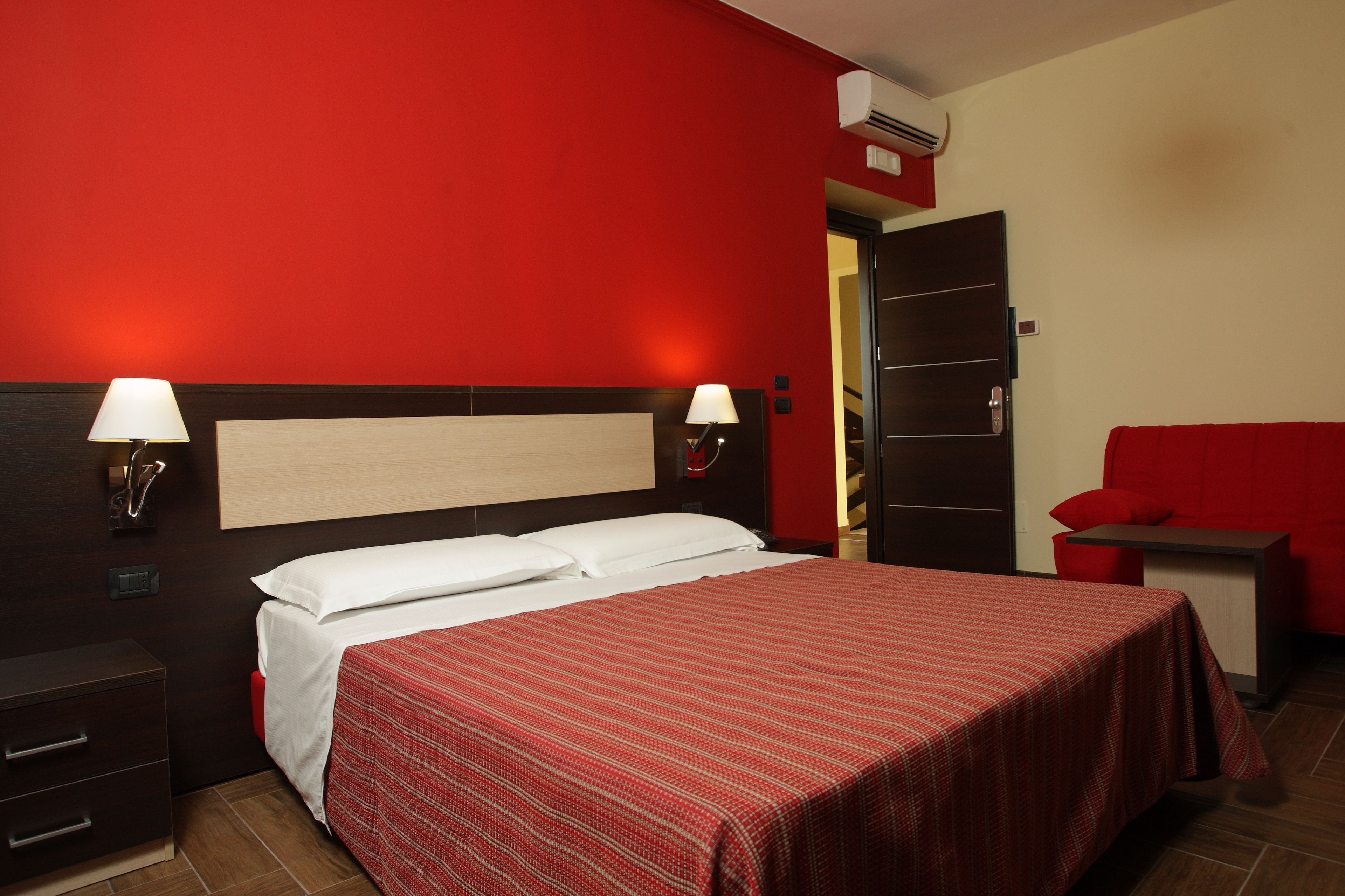 Habitación de hotel con una decoración en rojo y blanco, una cama con ropa blanca, dos lámparas de noche, una mesita de noche, una silla roja y un cabecero de madera.