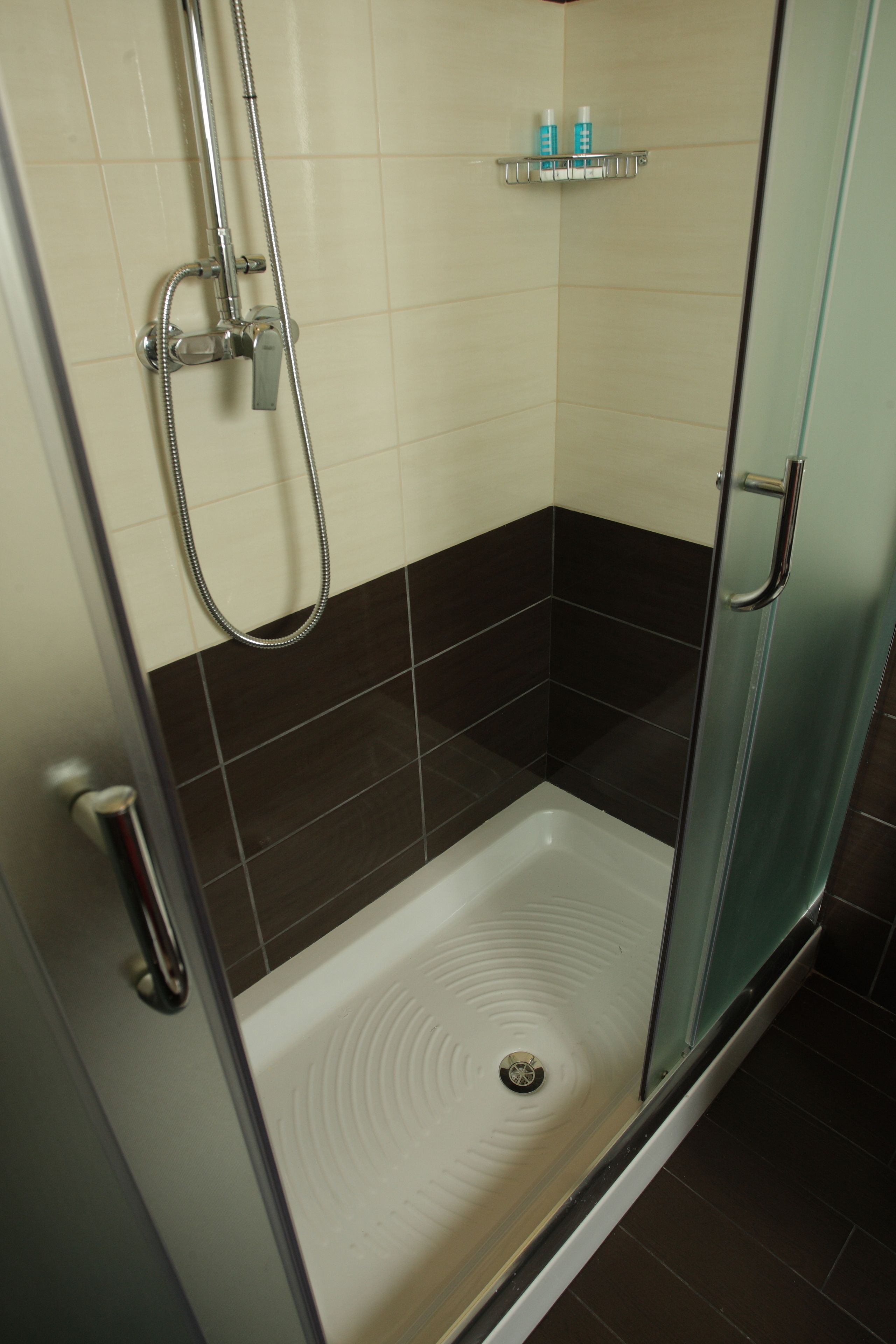 Une cabine de douche fermée par des parois en verre, avec un sol blanc, une douchette à main et une étagère au-dessus pour ranger les articles de toilette.