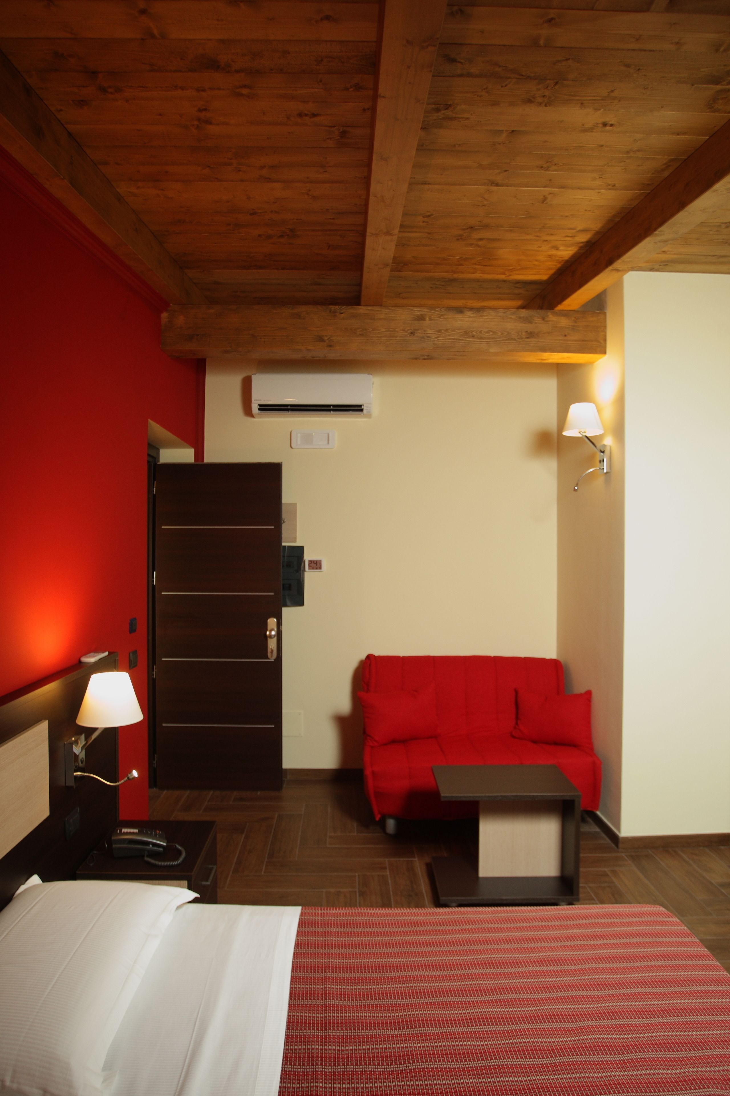 A hotel room with a wooden ceiling, a red wall, a bed with white bedding, a red sofa, a small table with a lamp, and an air conditioning unit.