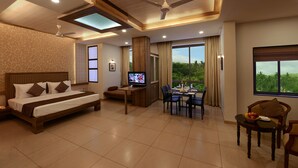 A modern hotel room with a large bed, a dining area, a TV, and a balcony with a view of greenery.