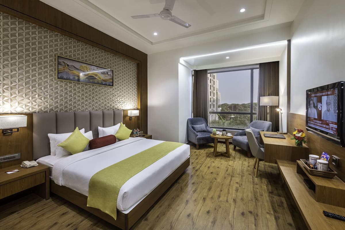 A hotel room with a bed, two armchairs, a desk with a TV, a ceiling fan, and a large window with a view.