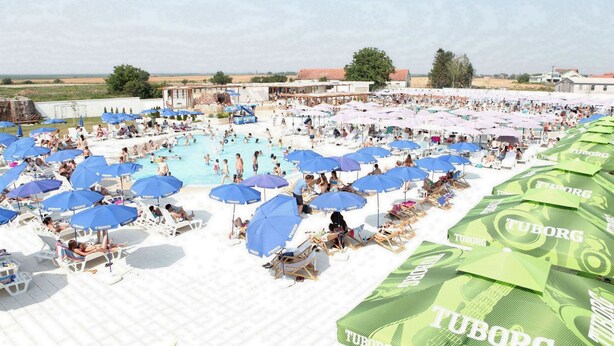 A pool area with multiple umbrellas and lounge chairs, surrounded by buildings and open fields. - Hotel Novella Uno (Novi Banovci)