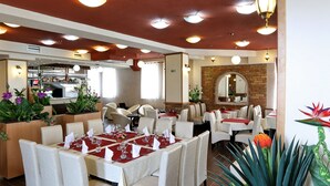 A restaurant with multiple dining tables set for guests, a kitchen area in the background, and a brick wall with a mirror. - Hotel Novella Uno (Novi Banovci)