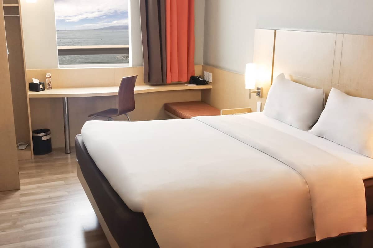 A hotel room with a bed, a desk, a chair, a lamp, and a view of the sea.