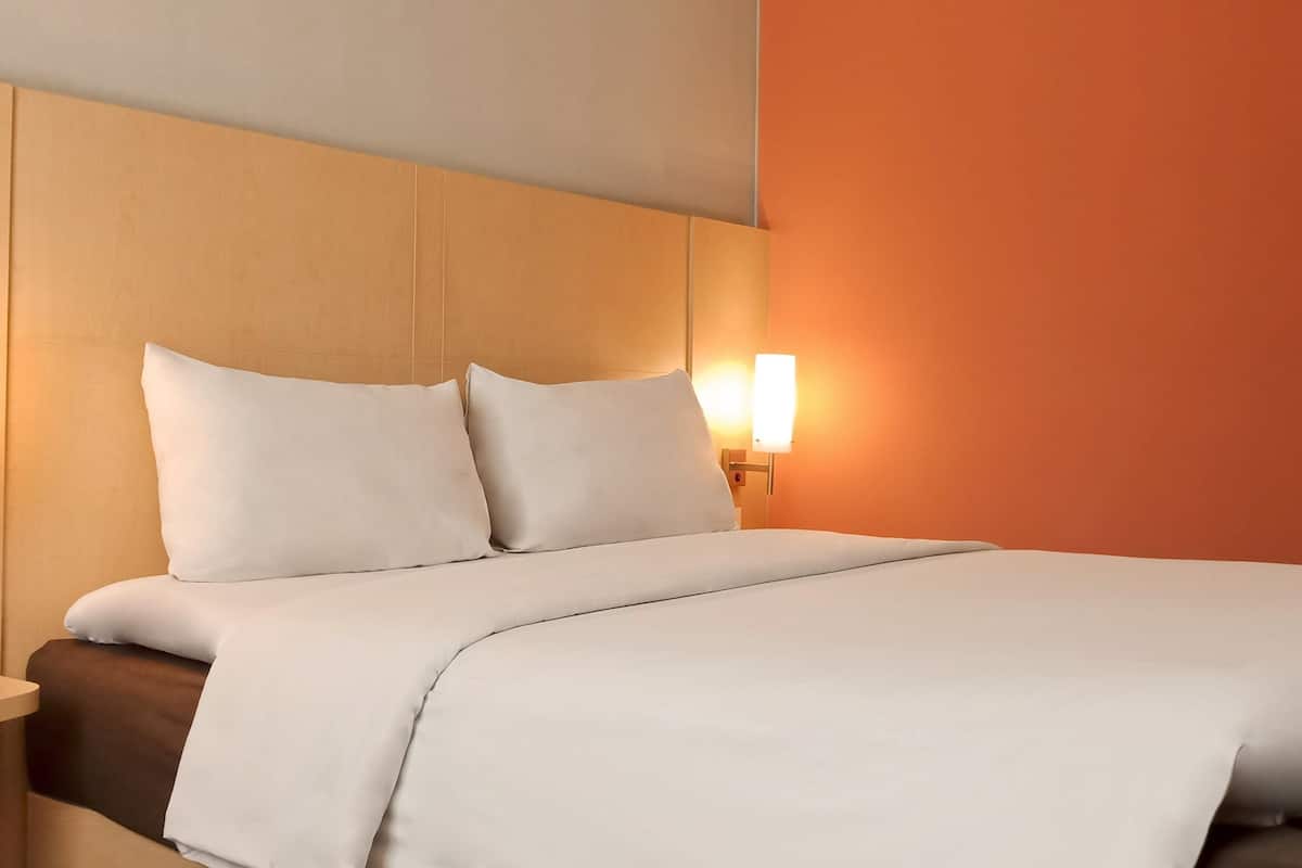 A hotel room with a bed, pillows, a nightstand, and a wall-mounted lamp.
