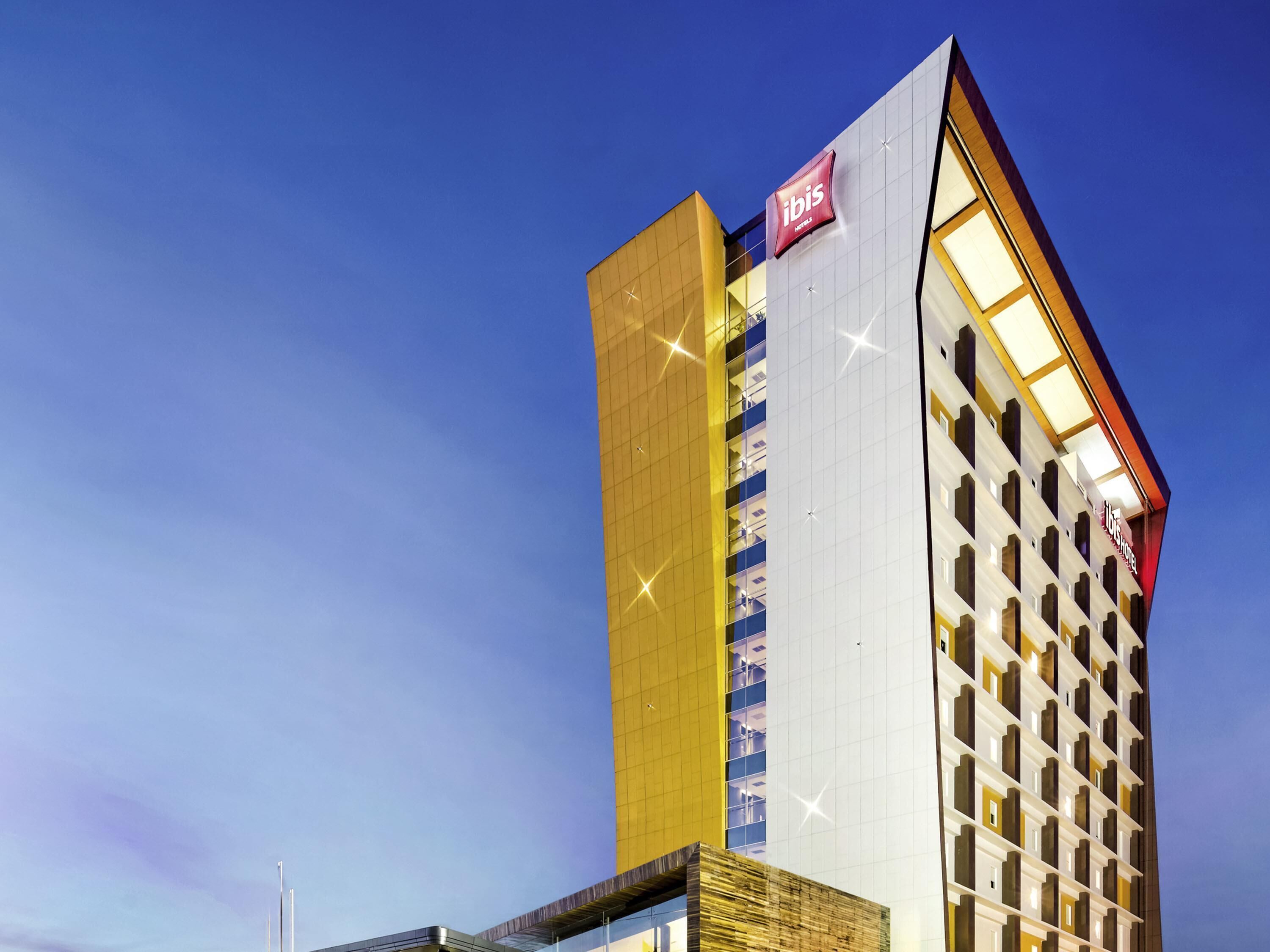 A modern hotel with a prominent 'ibis' sign, a tall structure with a mix of glass and solid panels, and a distinctive yellow and white facade.