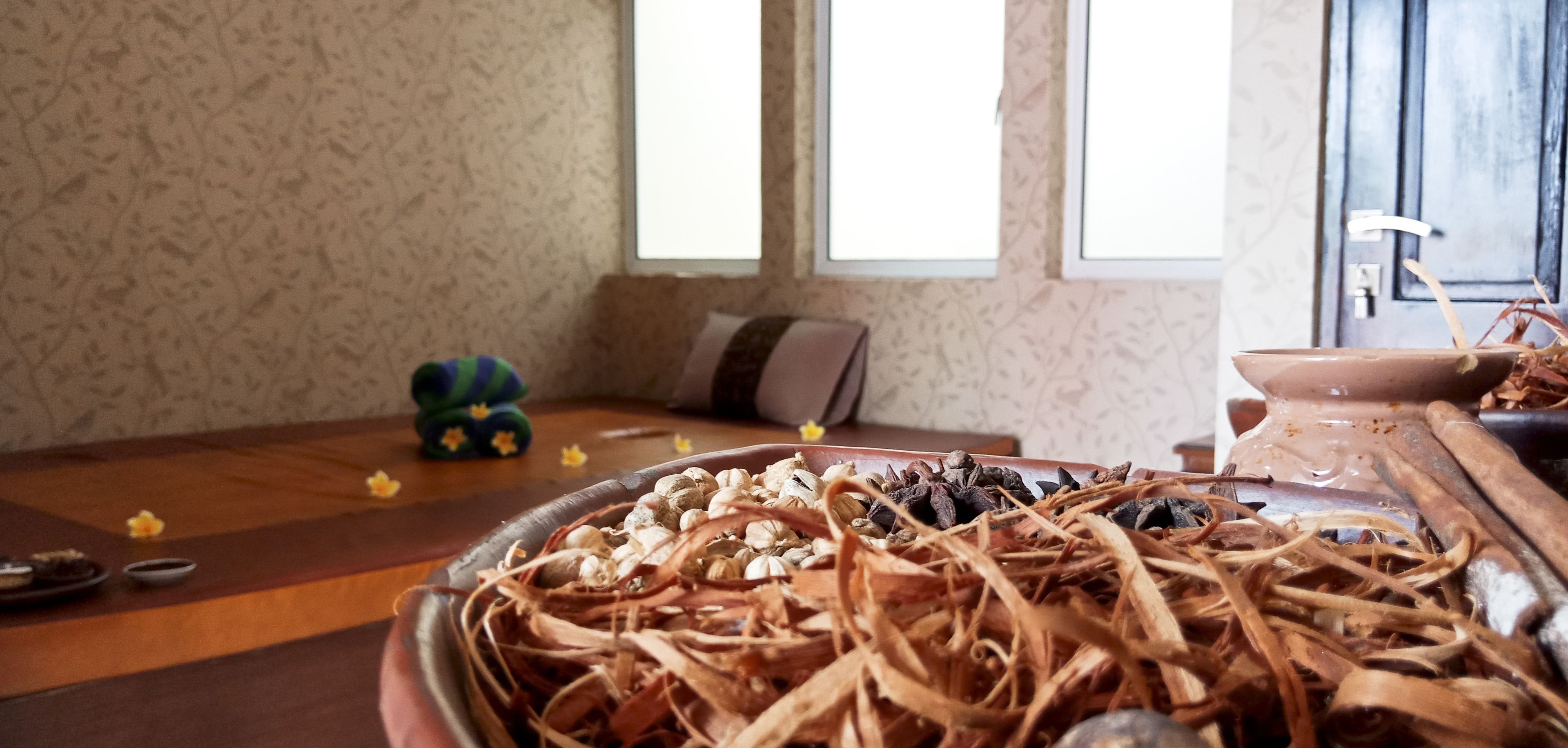 Ein Zimmer mit Holzboden, einer großen Schüssel voller verschiedener getrockneter Kräuter und Gewürze, einem kleinen Tisch mit Schüssel und Löffel sowie einem Fenster mit Vorhängen.