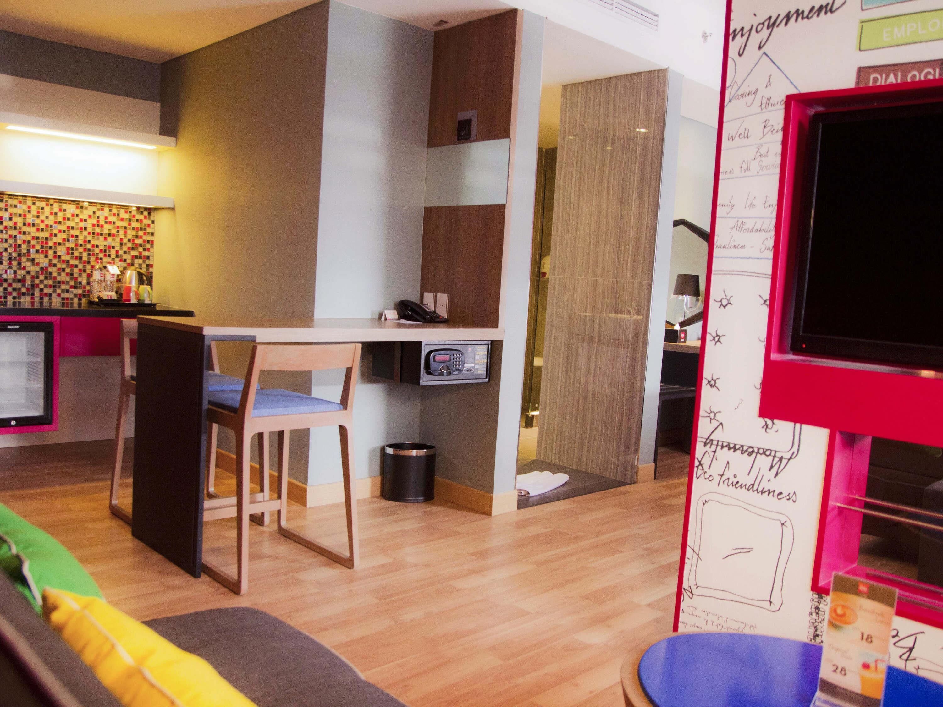 A modern hotel room with a kitchenette, a dining area, and a seating area with a sofa and chairs.