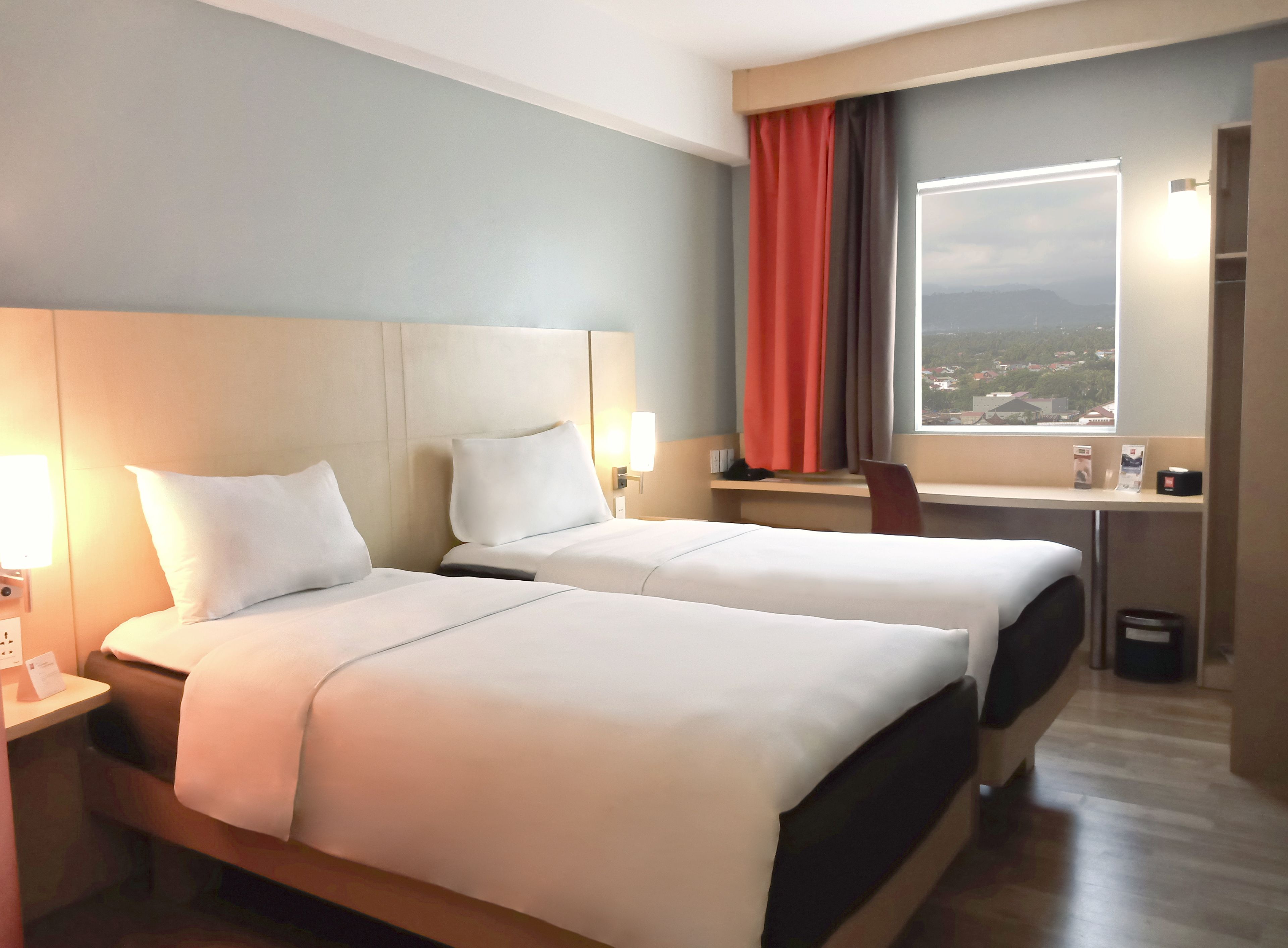 Habitación de hotel con dos camas, un escritorio, una silla, un ventanal con vistas y un cesto de basura.