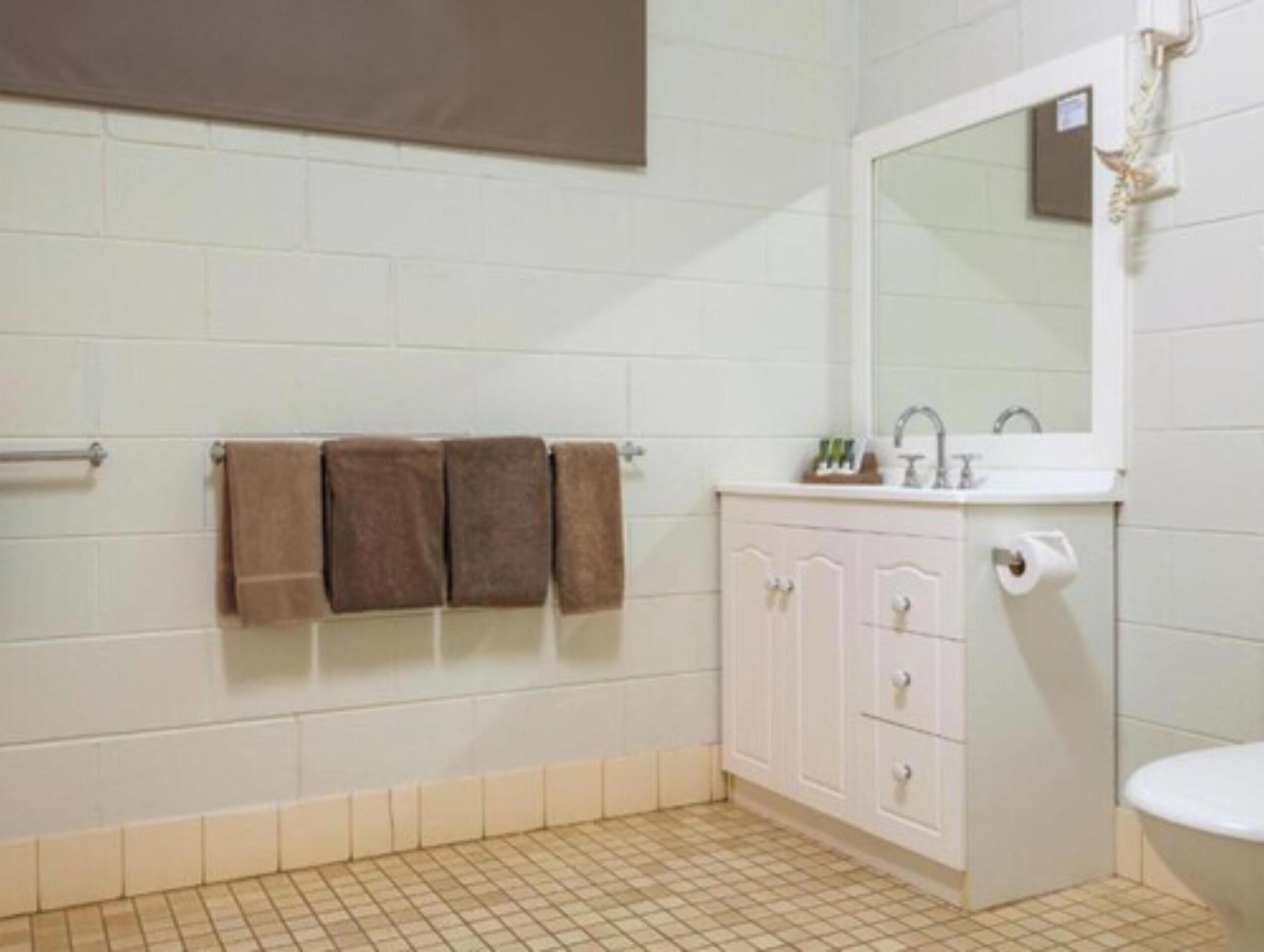 Une salle de bain avec des murs recouverts de carreaux blancs, un sol beige, un lavabo blanc avec un miroir et trois serviettes suspendues à un porte-serviettes.