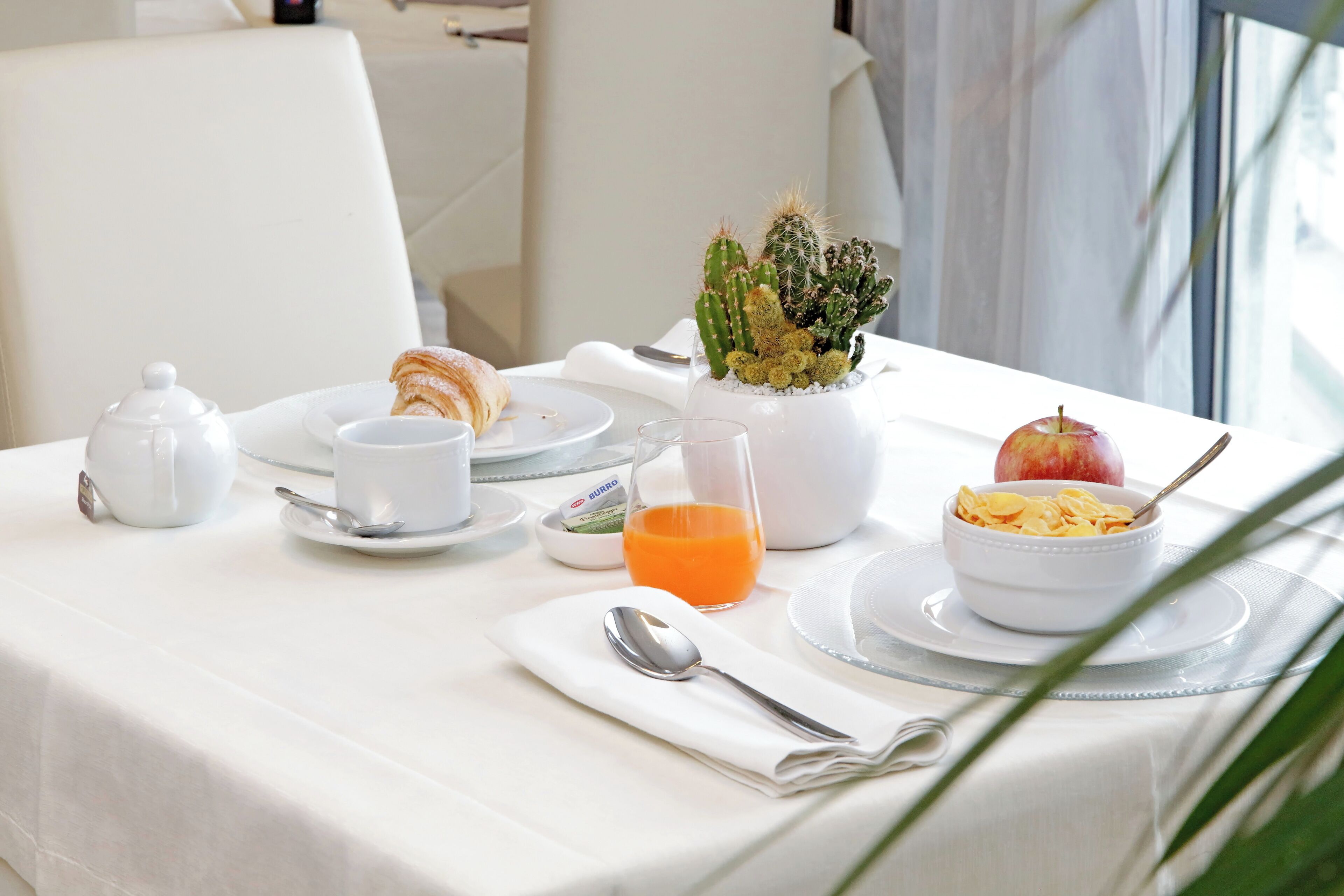 Taula de petit esmorzar amb estovell blanc, tetera, tassa, got de suc d'aranja, bol de cereals, croissant i planta de cactus.