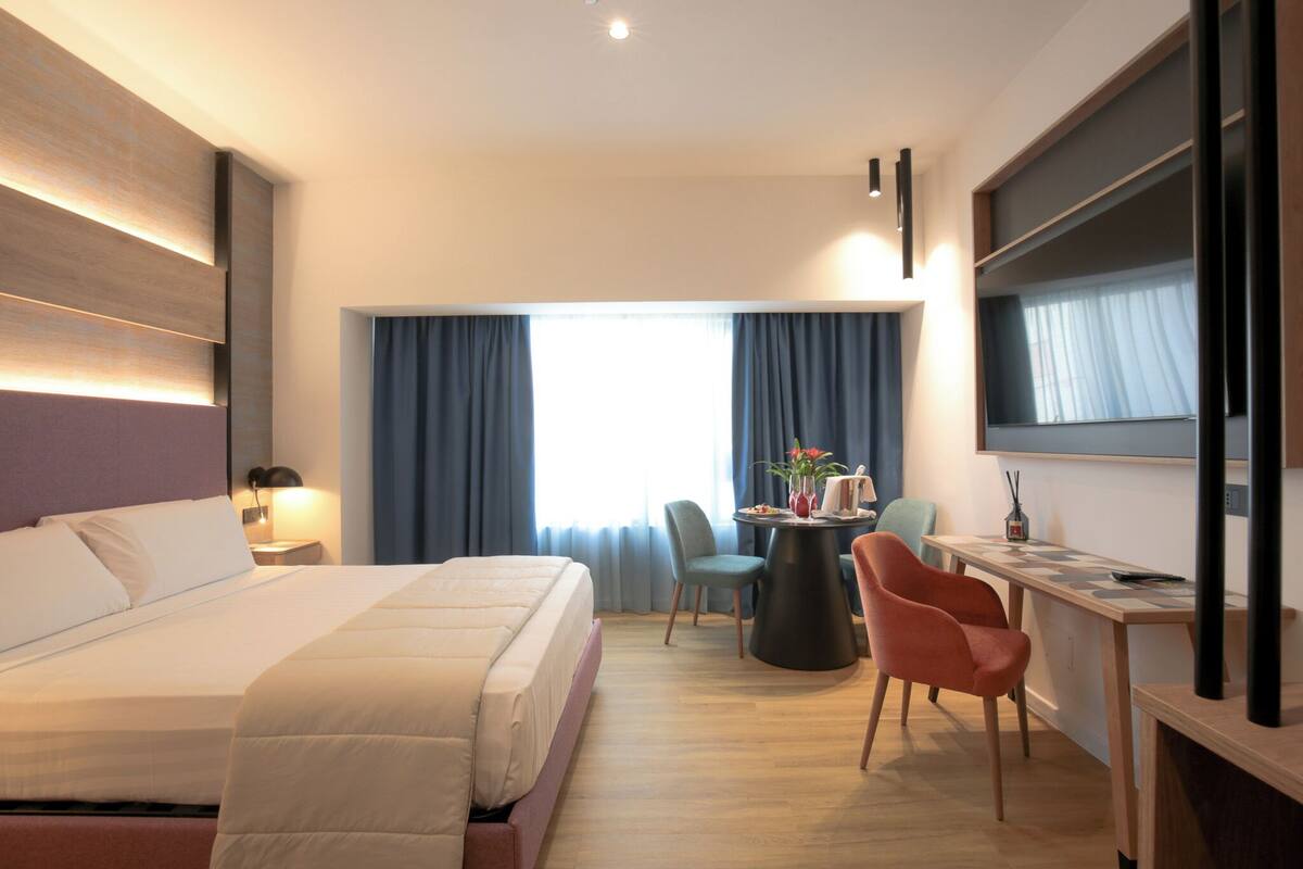 A hotel room with a large bed, a desk with a chair, a small table with a vase, and a large window with curtains.
