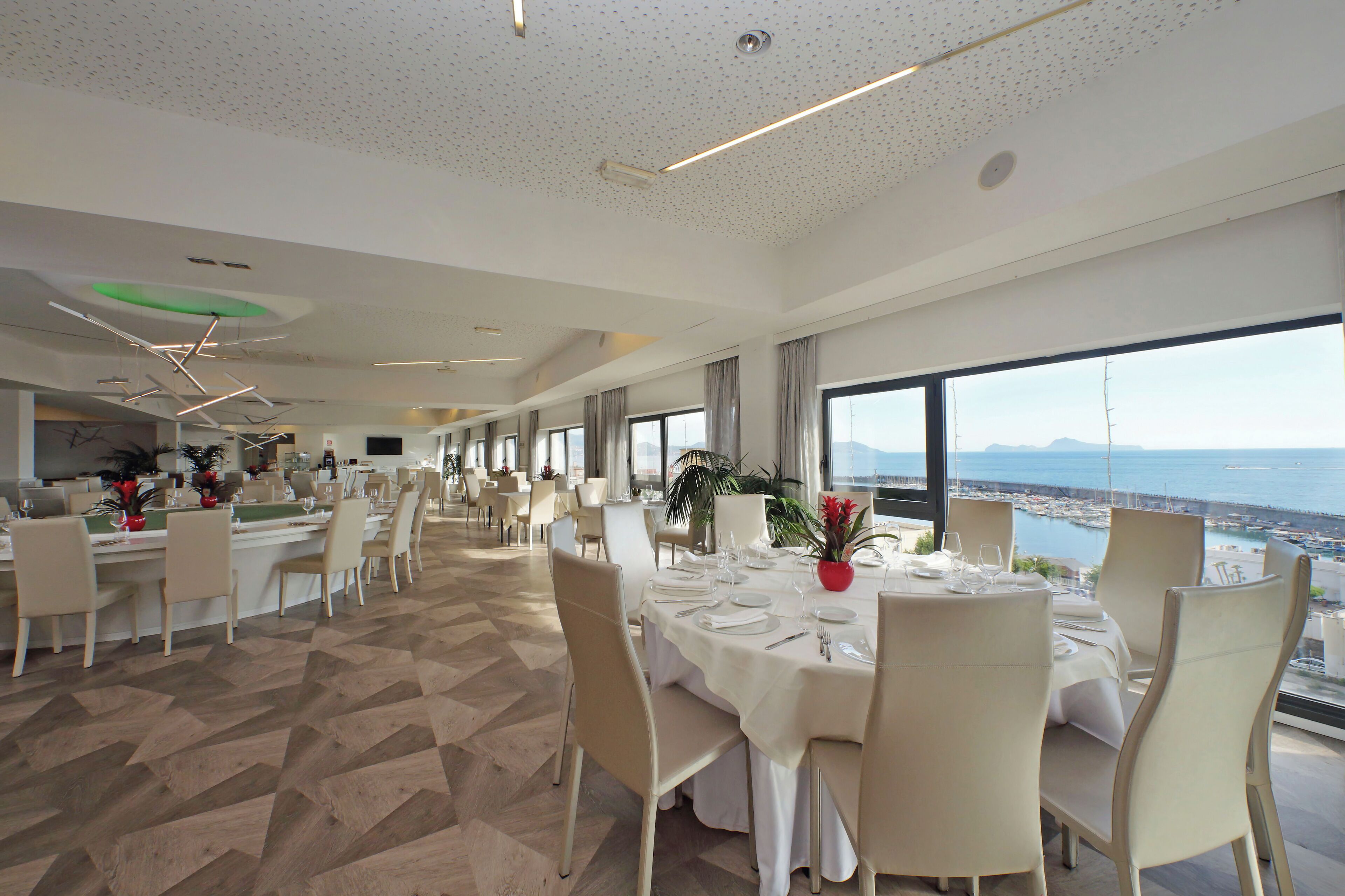 A spacious dining area with multiple tables set for dining, large windows offering a view of the sea, and a modern ceiling design.