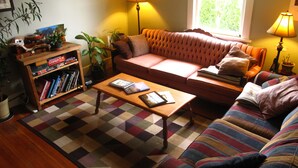 A cozy living room with a sofa, a coffee table, and a bookshelf. There are plants and lamps providing lighting. - Tofino Trek Inn (Tofino)