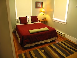 A neatly made bed with red bedding, a small bedside table with a lamp, and a striped rug on a wooden floor.