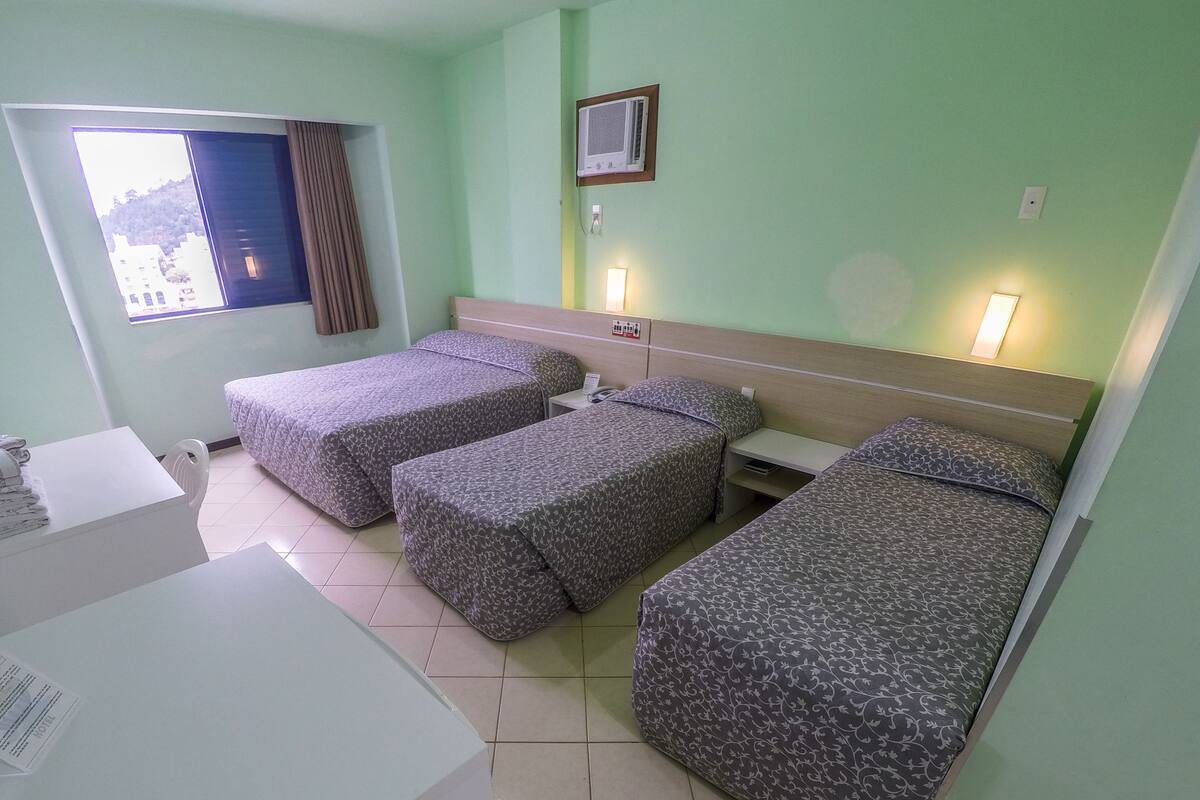 Een hotelkamer met twee eenpersoonsbedden, een bureau, een stoel, een raam met gordijnen en een luchtverkoeler aan de muur.
