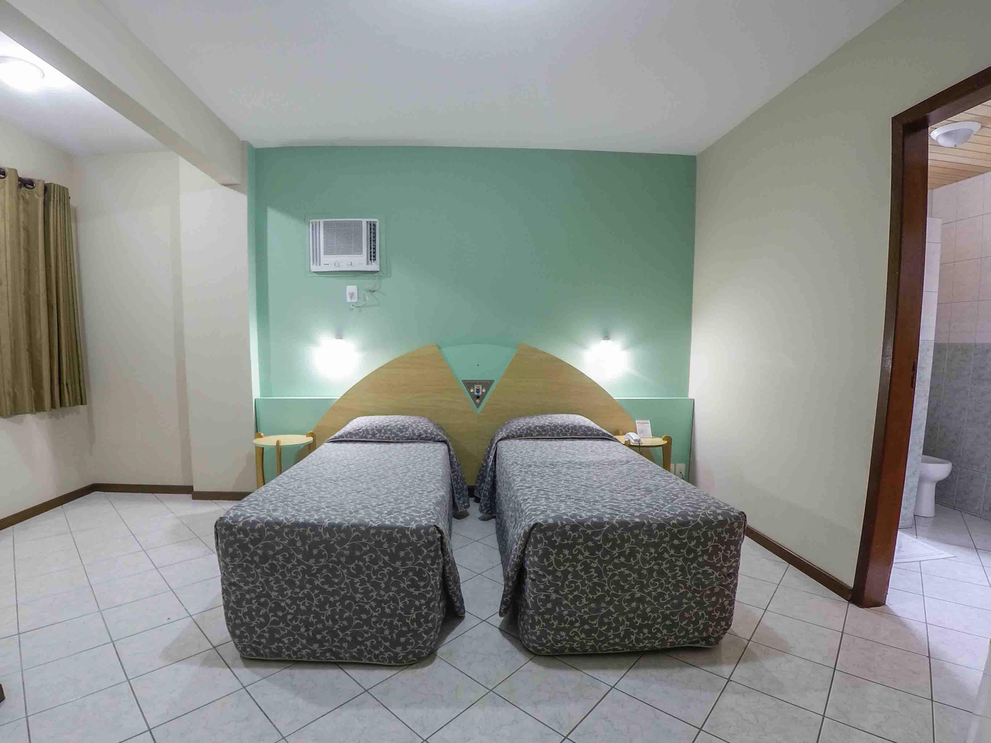 A room with two beds, a small table, a chair, an air conditioning unit, and a bathroom door.