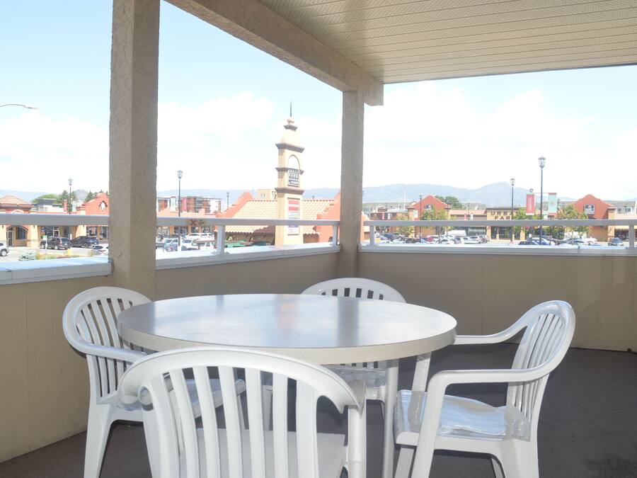 Un espacio al aire libre techado con una mesa redonda y cuatro sillas blancas, con vistas a un estacionamiento y edificios, incluyendo una torre a lo lejos.