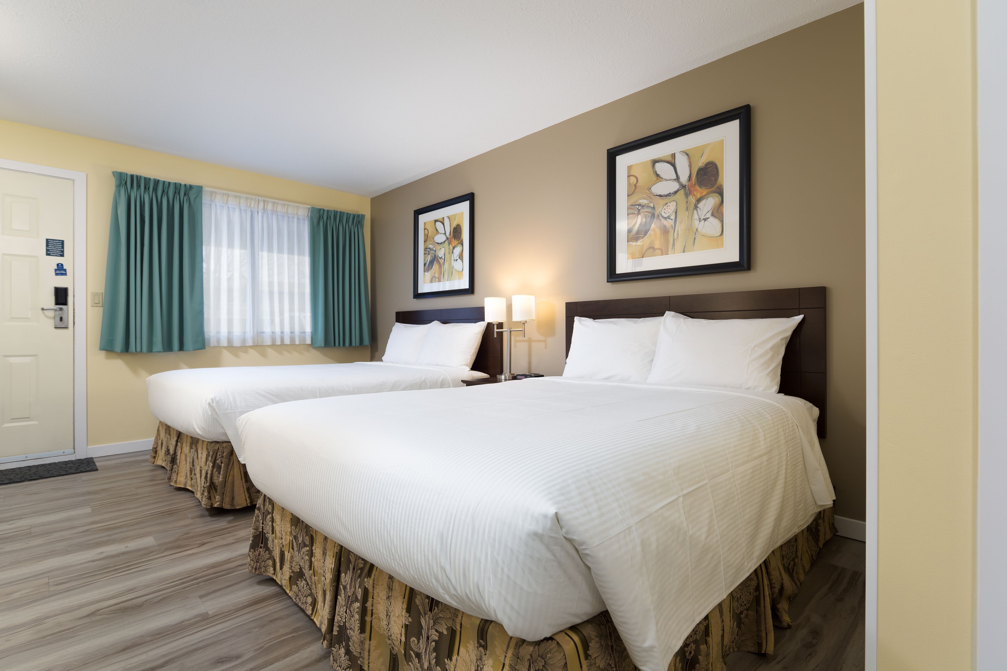 A hotel room with two beds, a window with curtains, a framed picture on the wall, and a nightstand with lamps.