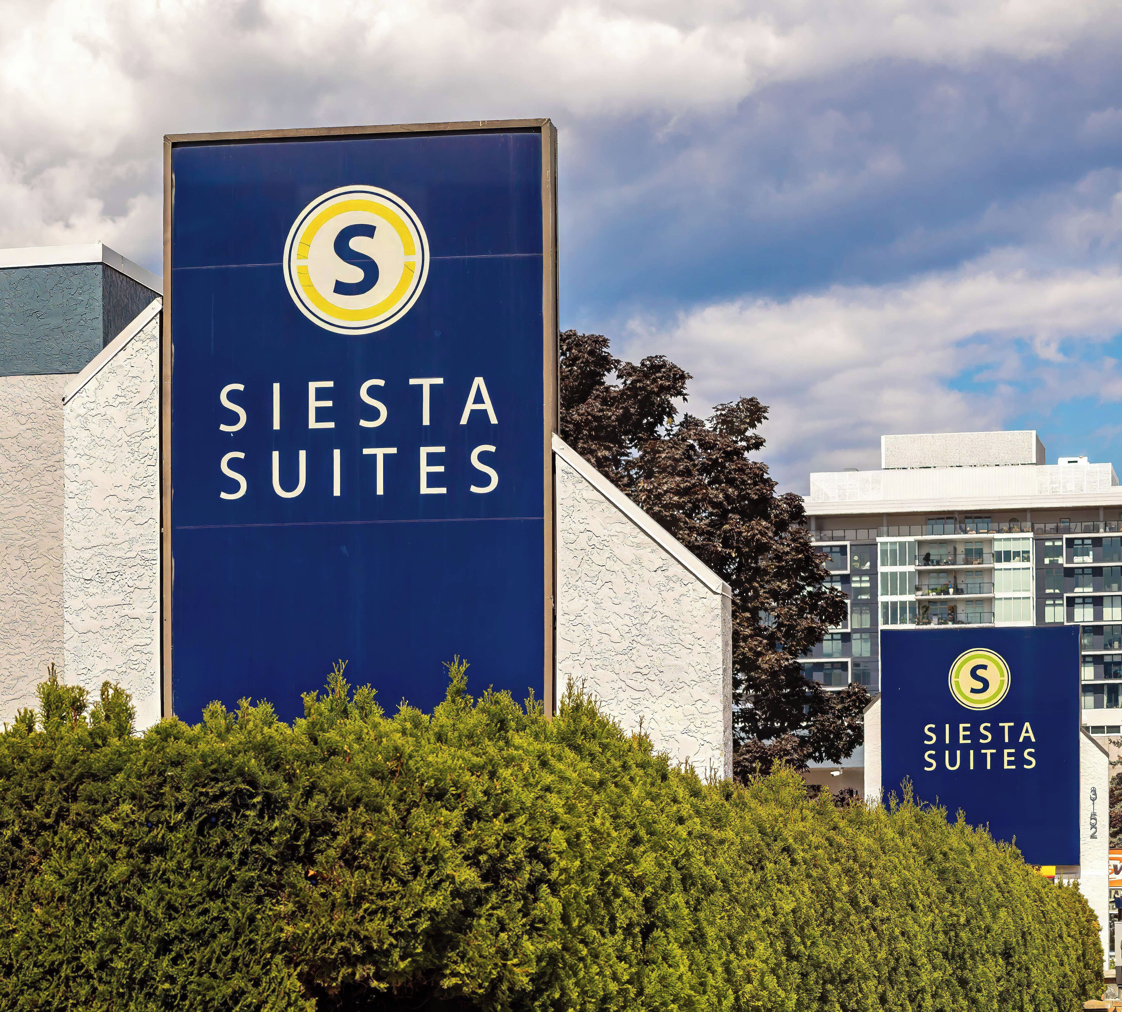 A large sign for Siesta Suites with a prominent logo, set against a backdrop of a building and trees.