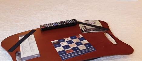A neatly made bed with white linens, a wooden tray with a remote control, a smartphone, and a magazine on the bed.