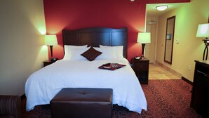 A hotel room with a large bed, two bedside lamps, a nightstand, a brown leather ottoman, and a red carpeted floor.