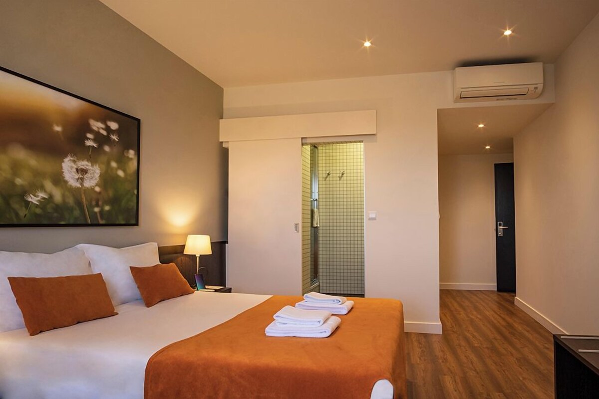 Habitación de hotel con cama, mesita de noche, lámpara, zona de ducha y unidad de aire acondicionado.