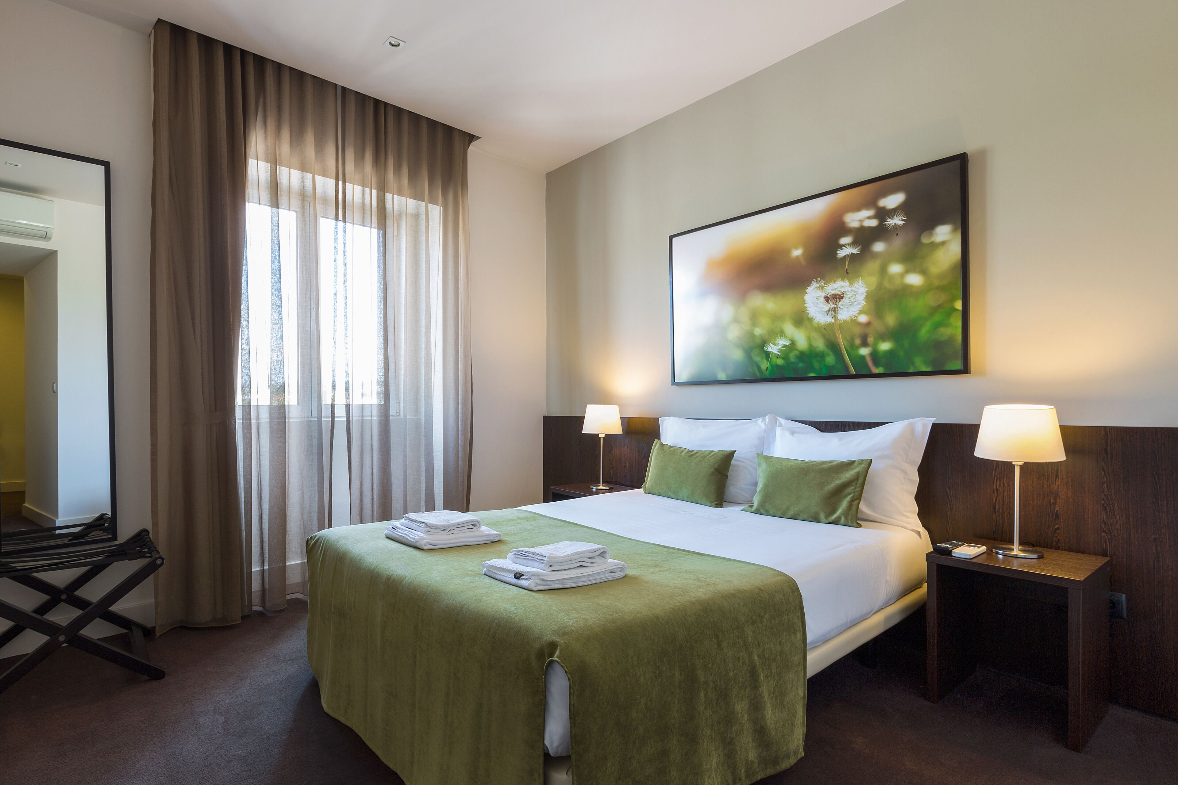 Quarto de hotel com uma cama grande, duas luminárias de cabeceira, um cobreleite verde, um quadro de flores na parede e uma janela com cortinas.