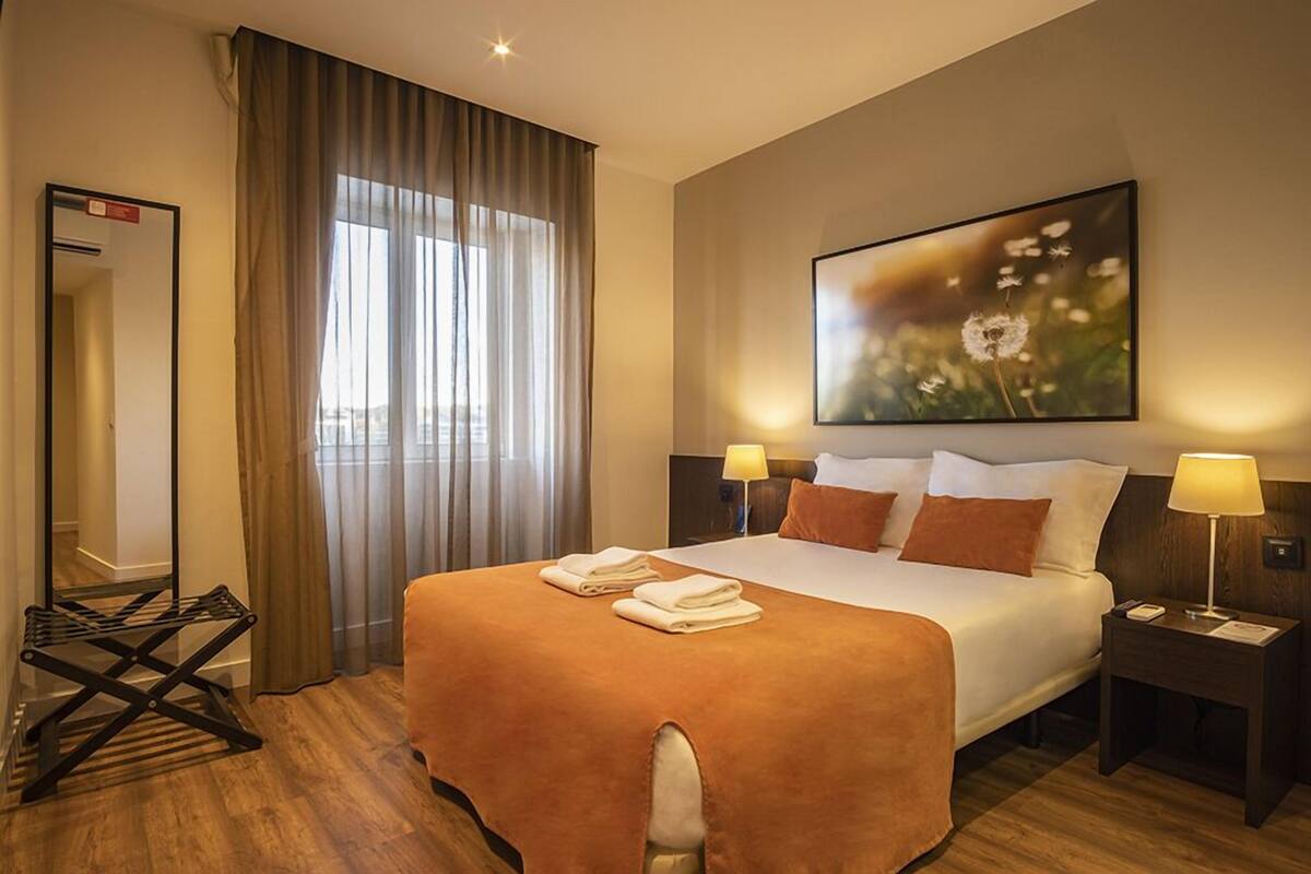 Una habitación de hotel con una cama grande, dos lámparas de noche, un espejo y un ventanal con cortinas.