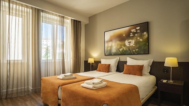 A hotel room with two beds, a large window with curtains, a painting on the wall, and bedside lamps. - Riversuites (Coimbra)