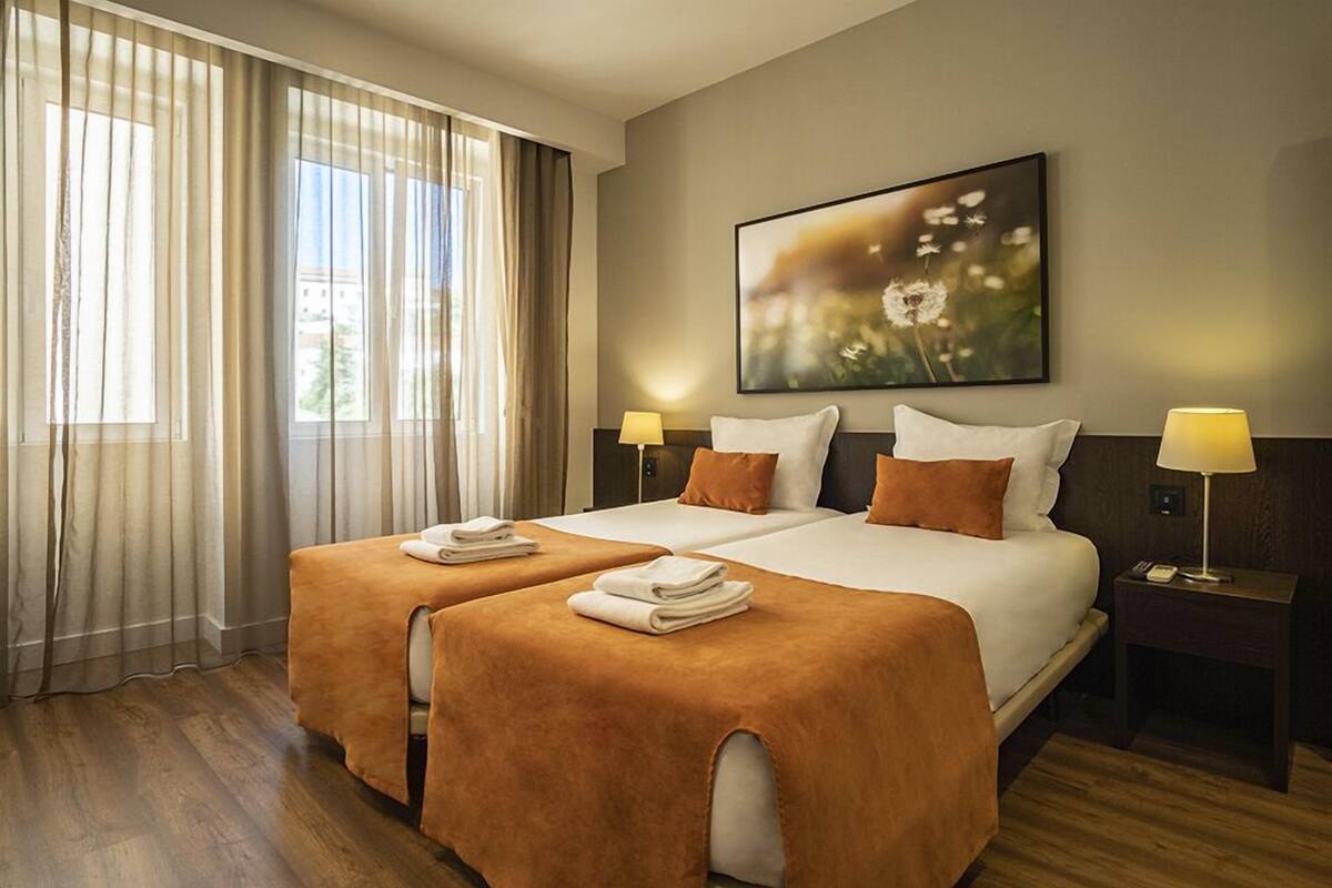 Habitación de hotel con dos camas, un ventanal con cortinas, un cuadro en la pared y lámparas de noche.