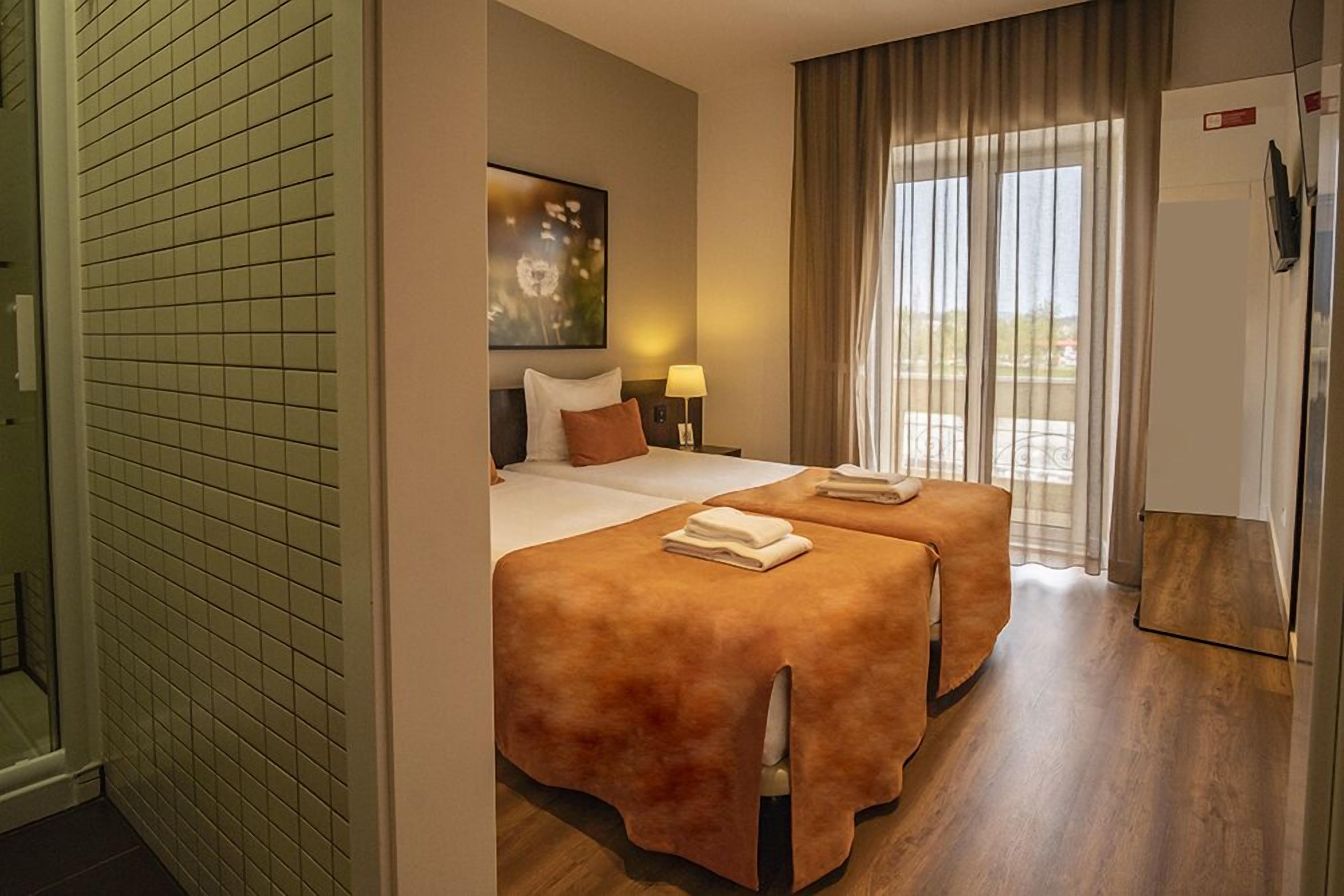 Ett hotellrum med två sängar, ett stort fönster med gardiner, en tavla på väggen och ett kylskåp.