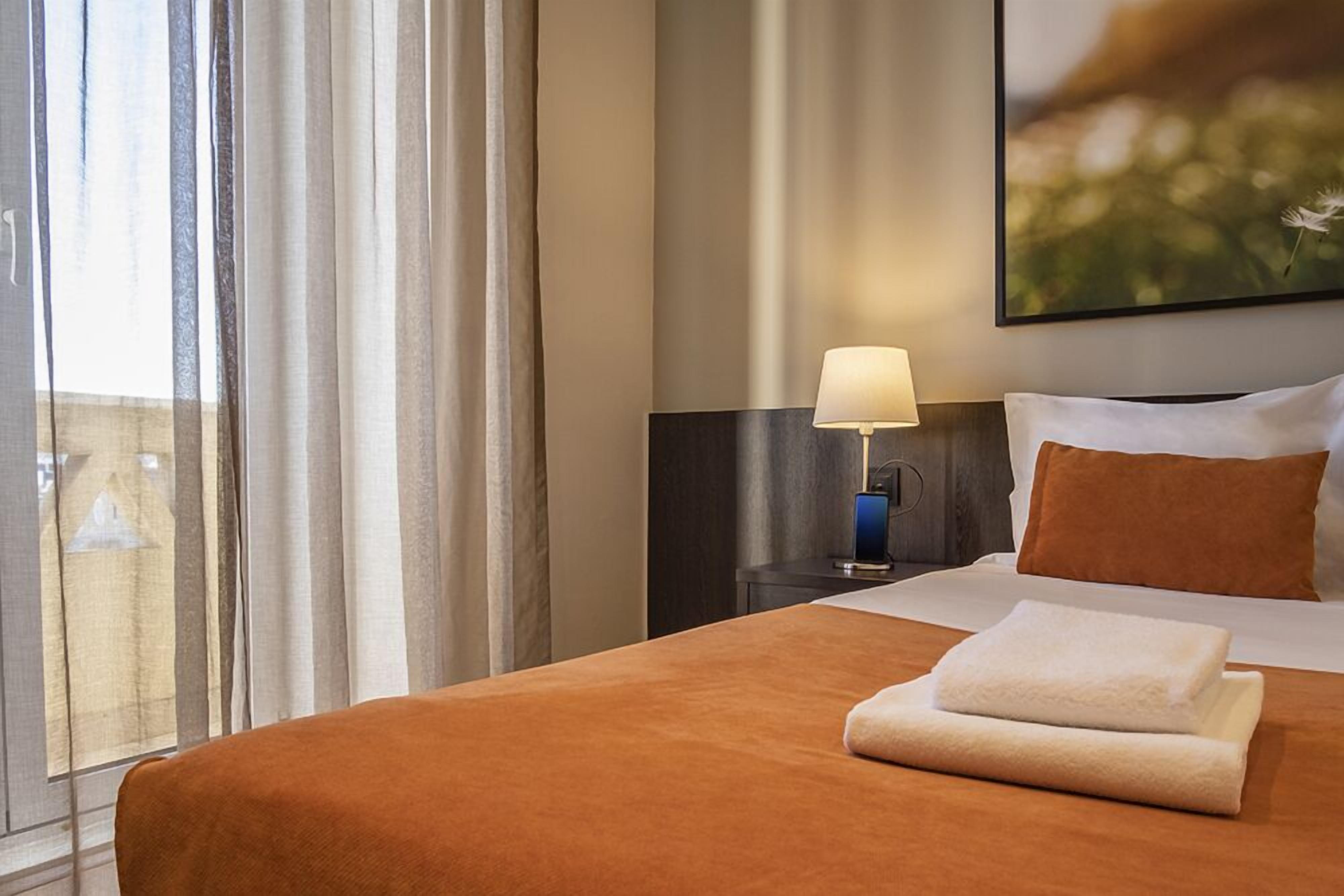 Una habitación de hotel con una cama grande, una mesita de noche con lámpara y una ventana con cortinas.