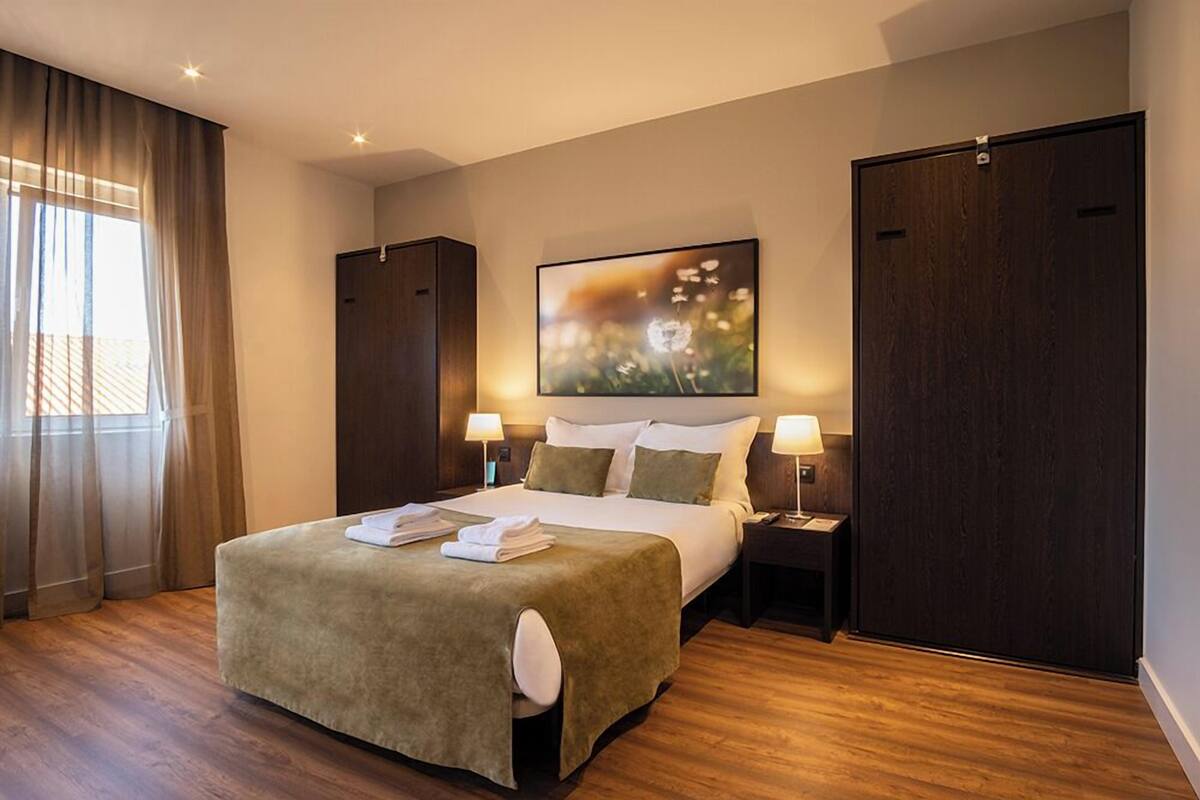Una habitación de hotel con una cama grande, dos mesitas de noche con lámparas, un armario y un ventanal con cortinas.