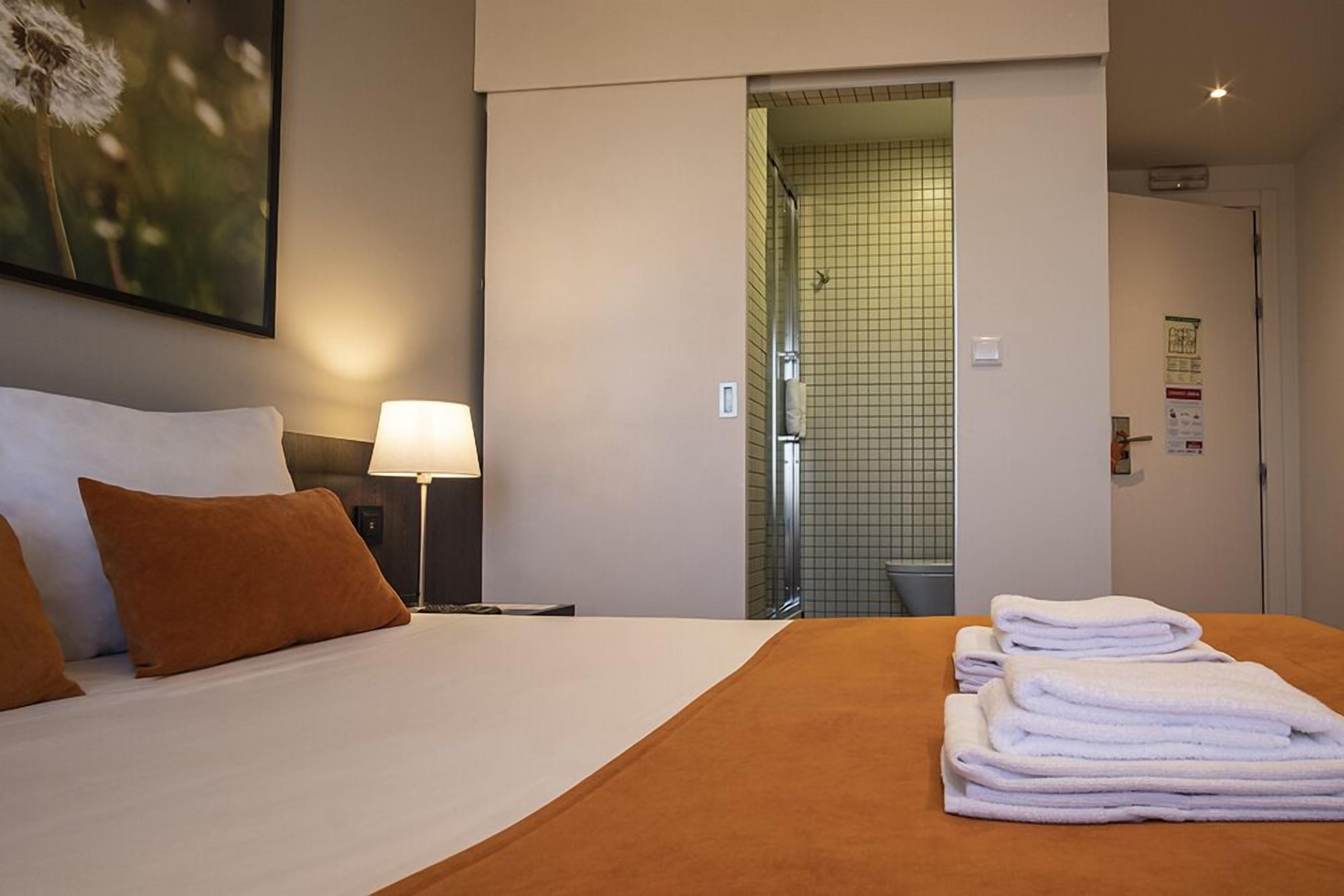 Una camera d'albergo con un letto, un comodino con una lampada, un bagno visibile attraverso una porta aperta e un mucchio di asciugamani sul letto.