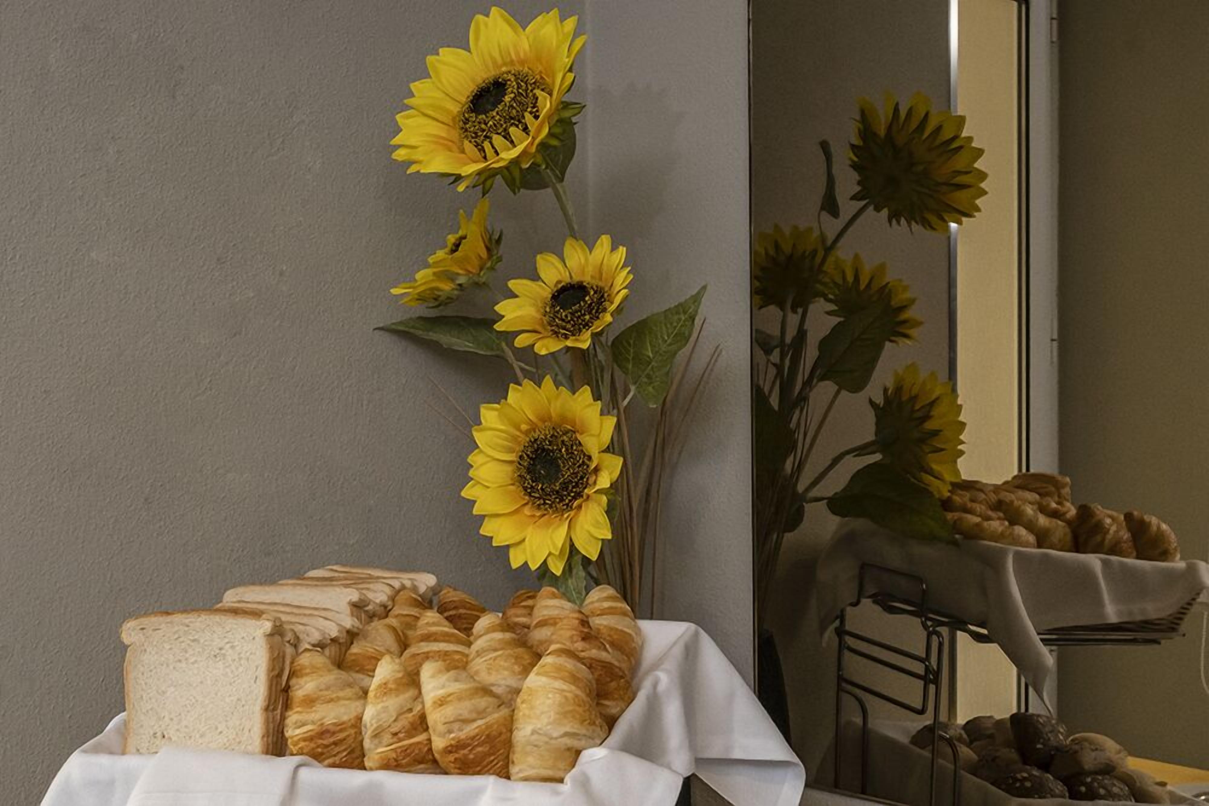 Un tavolo con pane e dolci, un vaso con girasoli e uno specchio che riflette la scena.