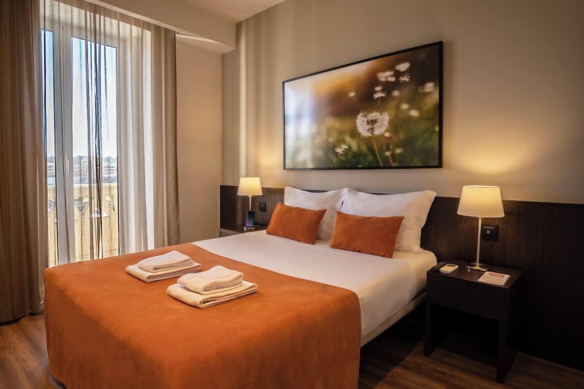 Habitación de hotel con una cama grande, dos lámparas de noche, un cuadro de diente de león y vistas a través de la ventana.
