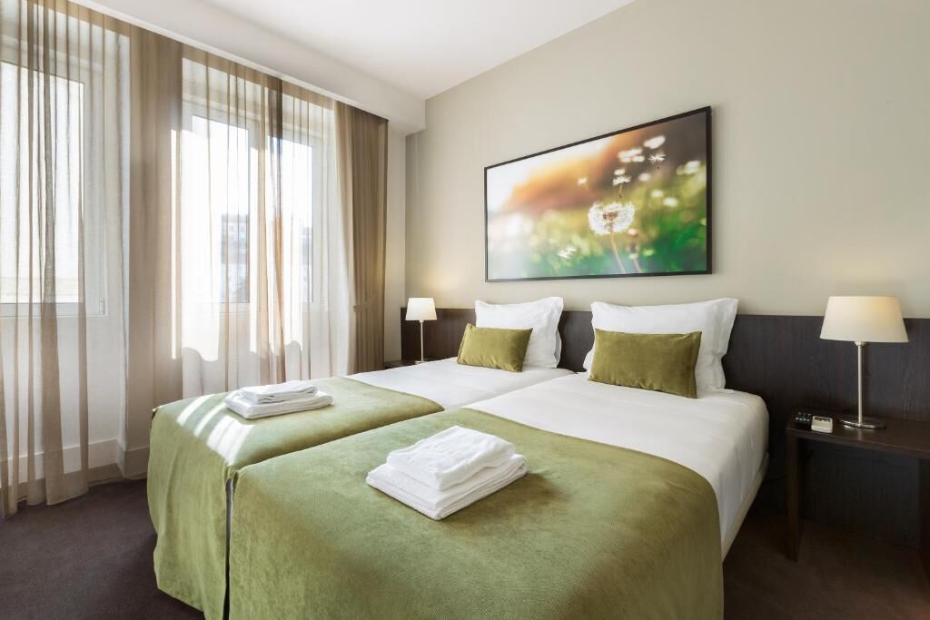 Habitación de hotel con dos camas, un ventanal con cortinas, un cuadro en la pared y una mesita de noche con lámpara.