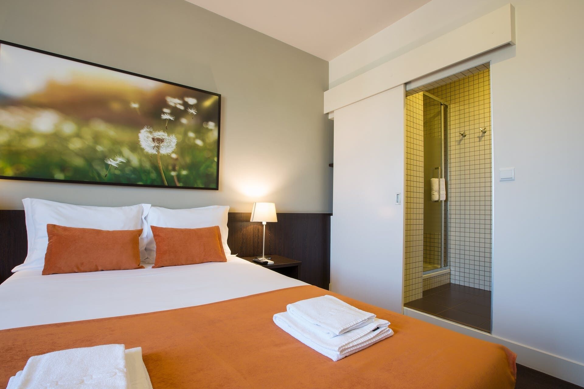 Habitación de hotel con cama, almohadas, lámpara, un cuadro y zona de ducha.